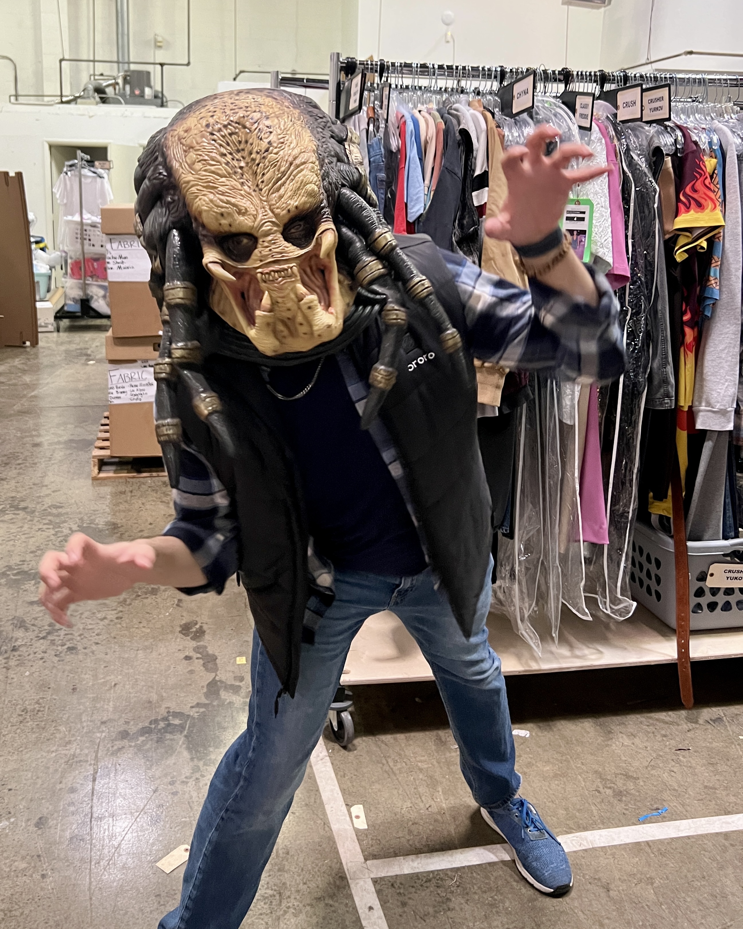 A Costumer wearing a scary mask with racks of costumes behind him.